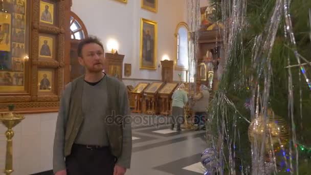 Un hombre barbudo mira un árbol decorado en la iglesia, con impresionantes iconos antiguos colgando de las paredes de un antiguo templo ortodoxo — Vídeo de stock