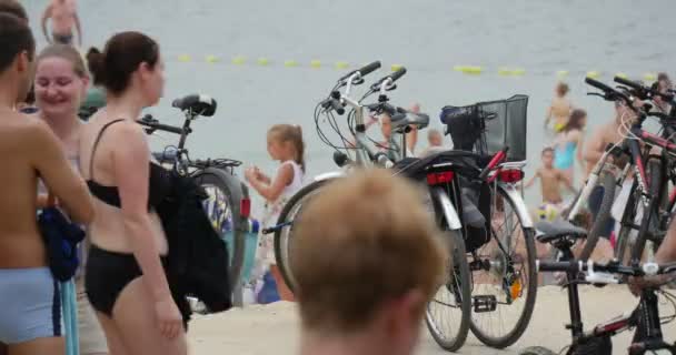 Adolescentes y personas de mediana edad en trajes de baño y uniformes deportivos Paseo y Gok a lo largo de la orilla del lago Bolko, con un montón de bicicletas en el fondo — Vídeo de stock