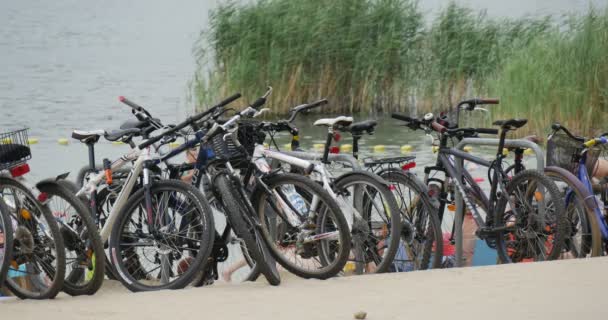 En hög med cyklar på stranden av sjön Bolko, människor fram och tillbaka och Reed växter som växer i bakgrunden i sommar — Stockvideo