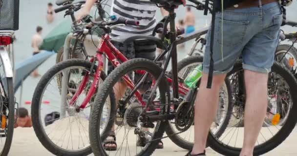 Una docena de bicicletas modernas en una orilla arenosa del lago Bolko, con un montón de jóvenes descansando en el fondo en verano — Vídeos de Stock