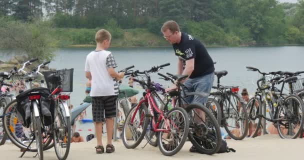 Padre Joven Ayuda a Su Hijo a Instalar Su Bicicleta y Ata Dos Bicicletas Juntos Mientras Están en una Arenosa Plage del Lago Bolko — Vídeos de Stock