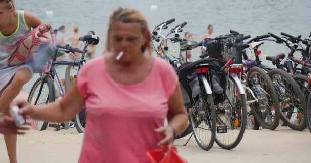Mujer comienza a fumar cuando los adolescentes montan bicicletas, prefiriendo la actividad deportiva en la orilla del lago Bolko en Polonia — Vídeo de stock