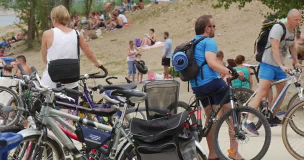 Hombre joven en ropa deportiva y gafas de sol se sienta en bicicleta y espera a alguien, mientras está entre la gente, que tienen un descanso en la Plage del lago Bolko — Vídeo de stock