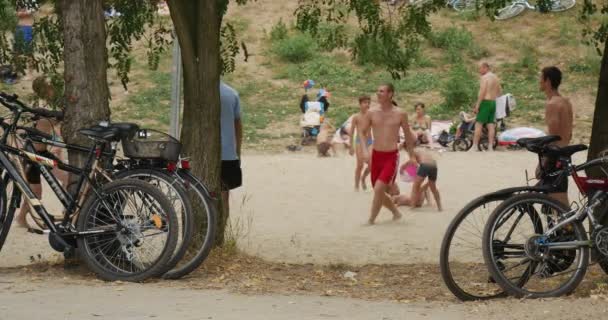 De jeunes garçons sportifs jouent au volley-ball sur une plage sablonneuse du lac avec plusieurs arbres verts et beaucoup de vélos et de touristes en journée, en été , — Video