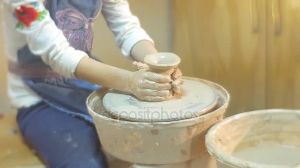 En un taller de producción de cerámica una pequeña chica rubia en blusa blanca y delantal azul hace una olla de arcilla y lo mira con entusiasmo — Vídeo de stock