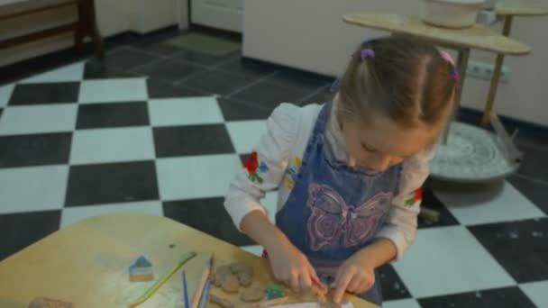 Niña rubia de seis años con ojos azules está trabajando en una mesa con muchos dibujos, estando dentro de un pequeño taller de cerámica, en interiores — Vídeos de Stock