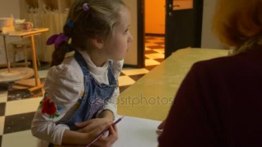 Küçük sarışın kız bir kare tablo ile bazı araçları oturuyor ve bir çanak çömlek Craft, beyaz Blose ve kot önlük, giyinmiş olmak öğrenme