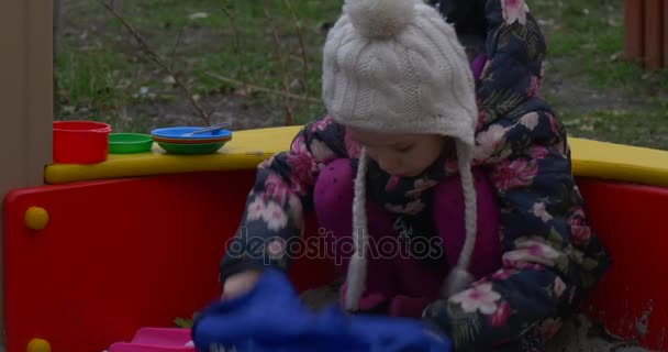 Una niña de seis años está jugando en un banco de color medio redondo para niños vestidos con un sombrero de punto blanco y un chaleco oscuro de color en otoño — Vídeos de Stock