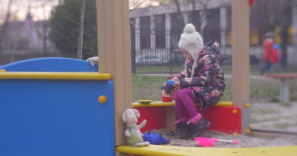 Шестилетняя блондинка играет на песочнице в парковой зоне в цветном пальто, вязаной шляпе с балабоном, розовые штаны, осенью в Польше — стоковое видео