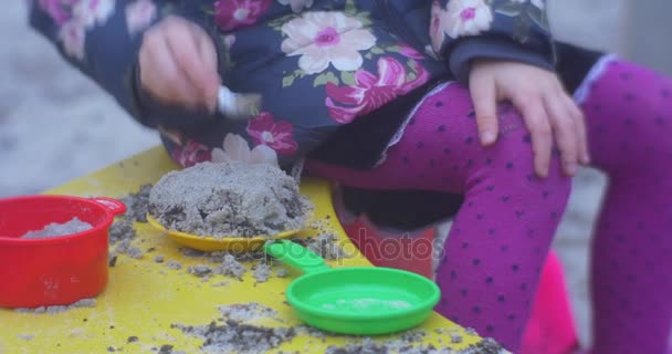 Spielplatz mit Sand und ein kleines Mädchen mit weißer Strickmütze und farbenfroher Weste unterhalten dort unweit eines Hauses im Herbst — Stockvideo