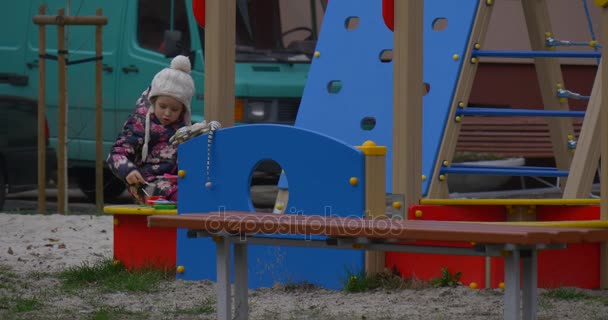 Spielplatz mit Sand und ein kleines Mädchen mit weißer Strickmütze und farbenfroher Weste unterhalten dort unweit eines Hauses im Herbst — Stockvideo