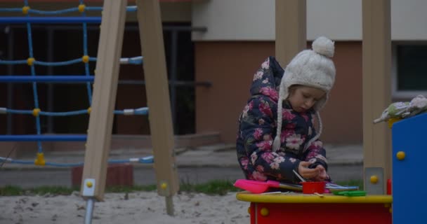 Gekleurde houten speeltuin met zand en een klein meisje in een witte gebreide muts en gekleurde Waistcoat Entertaing daar niet ver van een huis in de herfst — Stockvideo