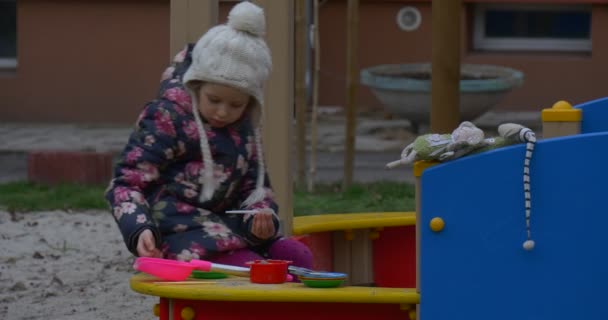 彩色木制操场与砂和一个小女孩在一个白色针织帽子和彩色的 Waiscoat Entertaing 那里不远的房子在秋天 — 图库视频影像