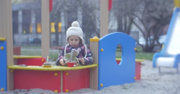 Spielplatz mit Sand und ein kleines Mädchen mit weißer Strickmütze und farbenfroher Weste unterhalten dort unweit eines Hauses im Herbst — Stockvideo