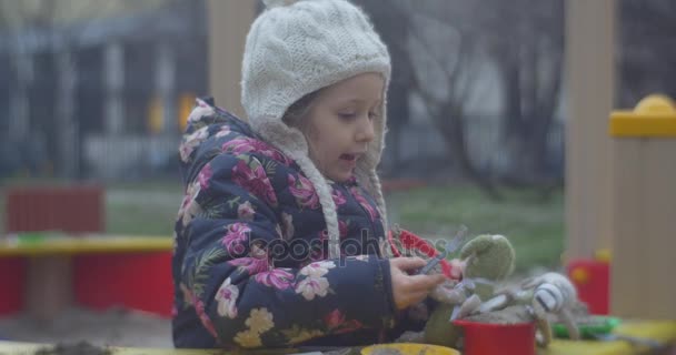 Ein kleines, sechs Jahre altes blondes Mädchen kocht in der Küche, während es im Herbst an einem bunten Sandkasten mit Plastikplatten, Löffeln und Töpfen steht. — Stockvideo