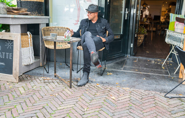 Бедняга в шляпе, одетый в черную одежду и ботинки, сидящий снаружи
