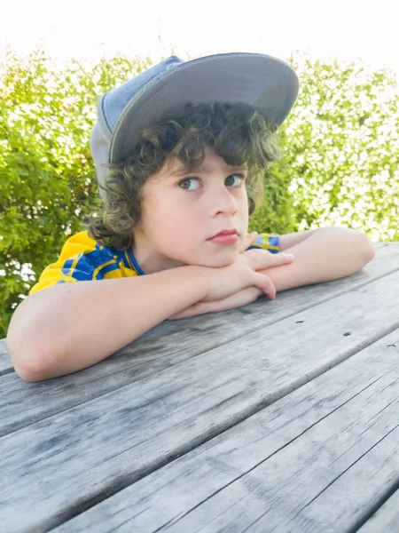 Pojke som bär stora cap på utanför tabellen vila hakan på armar — Stockfoto