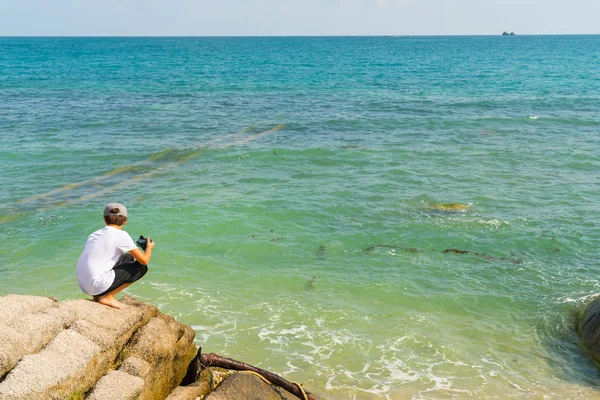 Menino segurando agachamentos câmera na rocha olhando para o mar em loc tropical — Fotografia de Stock