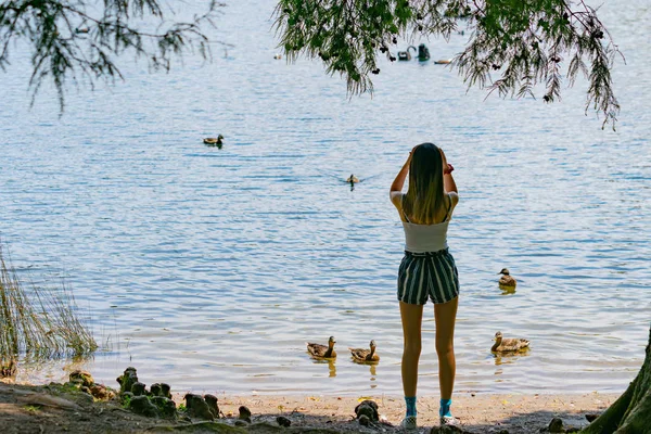 Teenage girl stands by lake edge with ducks in water