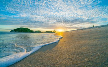 Güneş ufuktan yükselirken Moturiki adasıyla Maunganui Ana Kumsalı ve sıcak altın ve mavi renkli beyaz kabarık bulutlar..