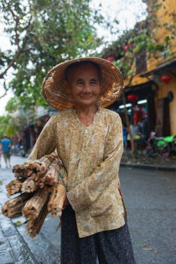 Hoi An Vietnam 17 Ekim 2020; Yüzü sert yaşam çizgileri gösteren yaşlı kadın yırtık pırtık bir koni şapka giyiyor ve bir sürü kamış taşıyor.