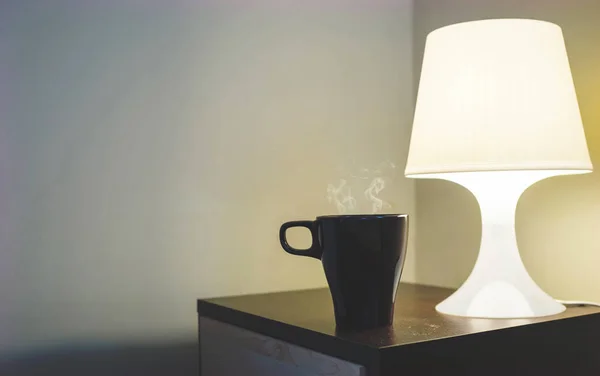 Tasse avec vapeur chaude et lampe sur la table d'armoire de lit de côté, matin — Photo