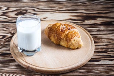 Bir bardak süt siyah bir zemin üzerine yakınındaki yuvarlak ahşap tabakta lezzetli bir kruvasan.