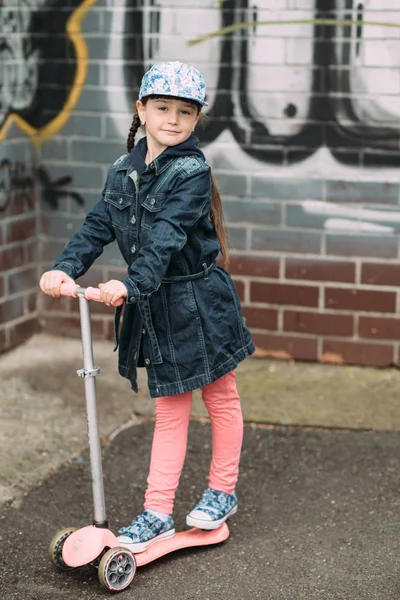 Linda niña en jeans cárdigan y en scooter en el fondo de la pared colorida urbana — Foto de Stock