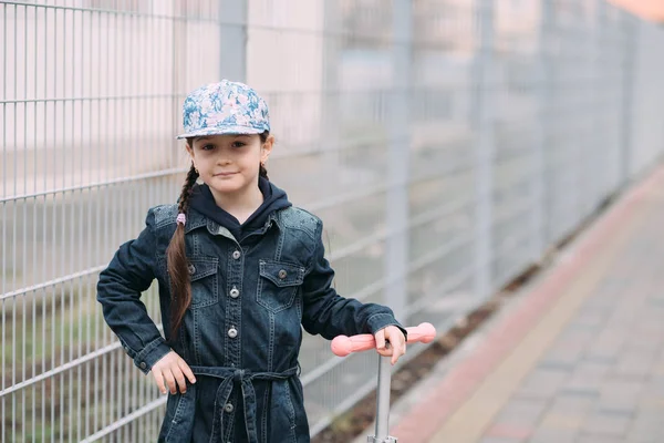 Linda niña en vaqueros cárdigan y en scooter en el fondo de la valla — Foto de Stock