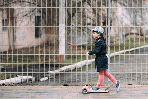 Милая маленькая девочка в джинсах и кардигане катается на скутере по улице — стоковое фото