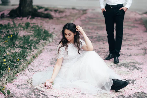 Retrato de una joven morena hermosa en vestido de novia blanco sentado en una hierba en un floreciente jardín de primavera . — Foto de Stock