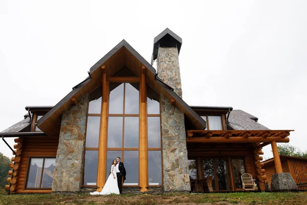 A woman in a wedding dress and a man in a stylish suit stand in front of a gorgeous house in the mountains