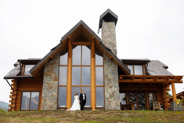 A woman in a wedding dress and a man in a stylish suit stand in front of a gorgeous house in the mountains