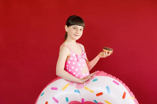 Tiro isolado de menina encantadora em maiô posando em estúdio com anel de borracha e segurando donut em sua mão. Menina bonita segurando círculo inflável de natação — Fotografia de Stock