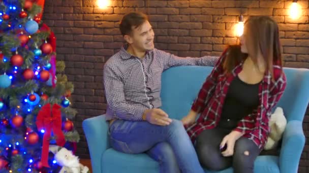 Молода пара сидить на дивані в новорічному інтер'єрі. Чоловік ніжно обіймає дружину — стокове відео