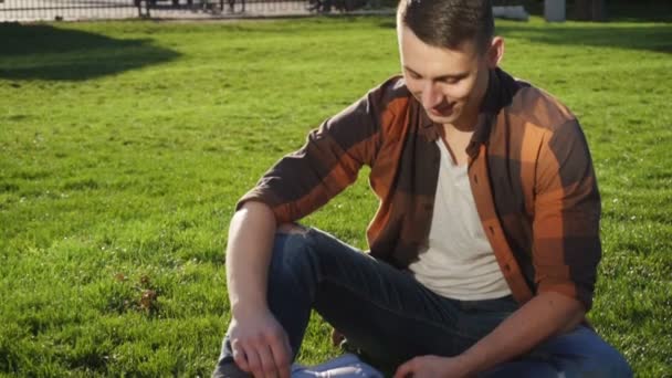 Молодой человек сидит на траве его девушка пришла и закрыть глаза сюрприз — стоковое видео