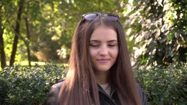 Mujer joven sentada en un banco del parque y sonriendo a la cámara en cámara lenta — Vídeo de stock