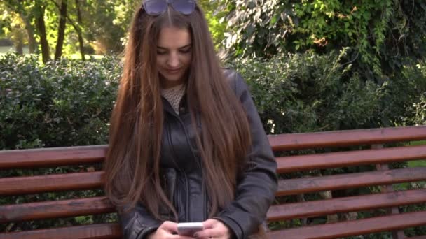Beautiful woman with long hair uses cell smartphone outdoors in the park — ストック動画