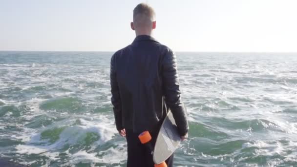 Молодой человек в черной кожаной куртке держит свою длинную доску у моря — стоковое видео