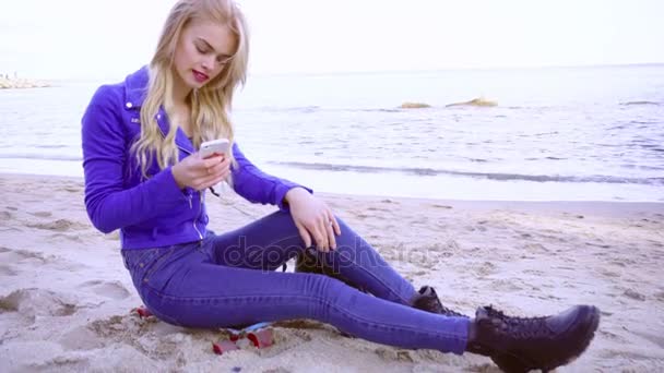 Chica rubia joven se sienta en la arena en la orilla del mar con su teléfono — Vídeos de Stock