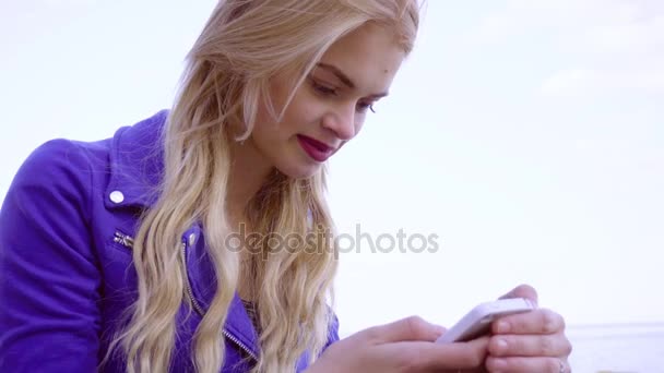 Menina loira jovem senta-se na areia na costa do mar com seu telefone — Vídeo de Stock