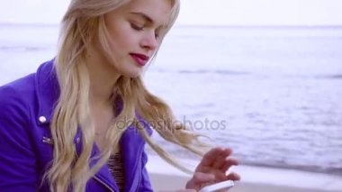 Genç sarışın kız telefonuyla ilgili kum deniz sahilinde oturur