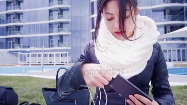 Ung brunett kvinna sitter utanför och lyssnar på musik i hörlurar — Stockvideo