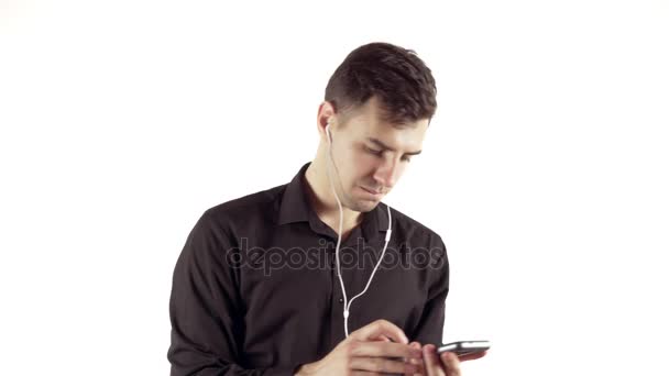 Porträtt av ung stilig man i svart skjorta och hörlurar Lyssna på musik på sin telefon som isolerad på en vit bakgrund — Stockvideo