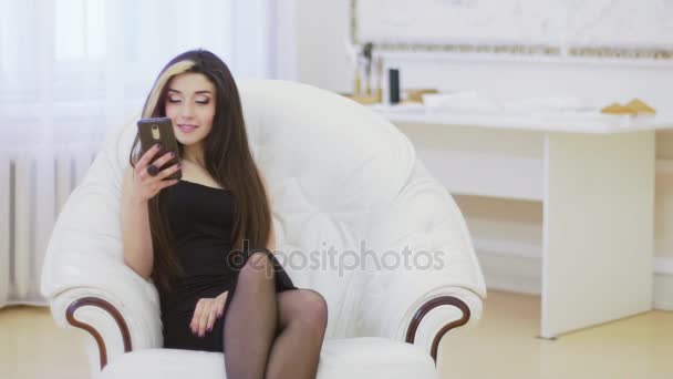 Schöne junge Model-Frau posiert im Studio auf einem weißen Sessel mit ihrem Smartphone — Stockvideo