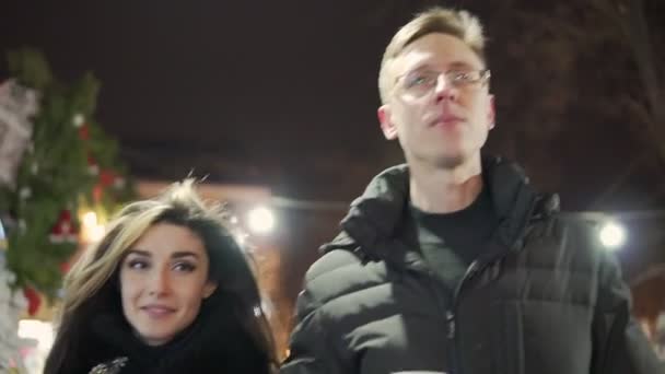 Casal jovem andar togerher perto de luzes de Natal no mercado de Natal com uma xícara de chá quente — Vídeo de Stock