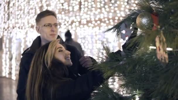 Feliz pareja atractiva de pie cerca del árbol de Año Nuevo en un mercado de Navidad por la noche. Hermosas luces de fondo . — Vídeo de stock