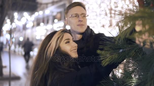 Счастливая привлекательная пара, стоящая рядом с новогодней елкой на рождественском рынке ночью. Красивый фон . — стоковое видео