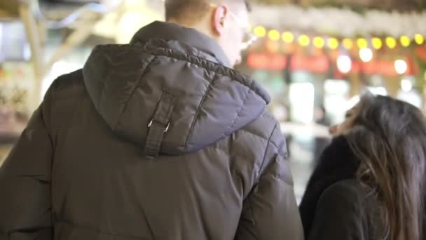 Pareja joven camina togerher cerca de las luces de Navidad en el mercado de Navidad. Vista trasera — Vídeos de Stock