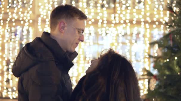Feliz pareja atractiva bailando cerca del árbol de Año Nuevo en un mercado de Navidad por la noche. Hermosas luces de fondo . — Vídeos de Stock